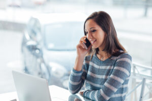 A woman is on the phone and sitting at her computer. She is wearing a blue and white-striped sweater and is having a pleasant conversation on the phone.