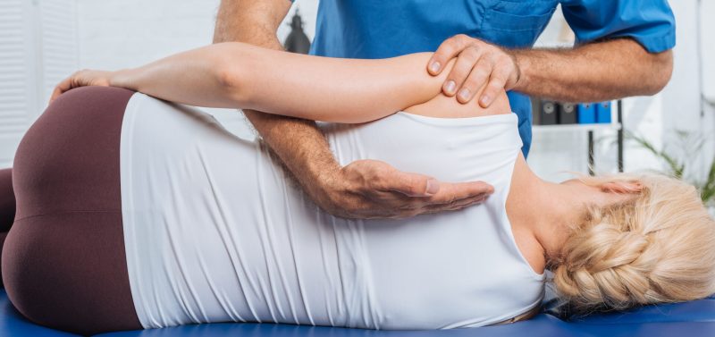 A chiropractor makes an adjustment on his patient. He is helping her around the upper part of her back.